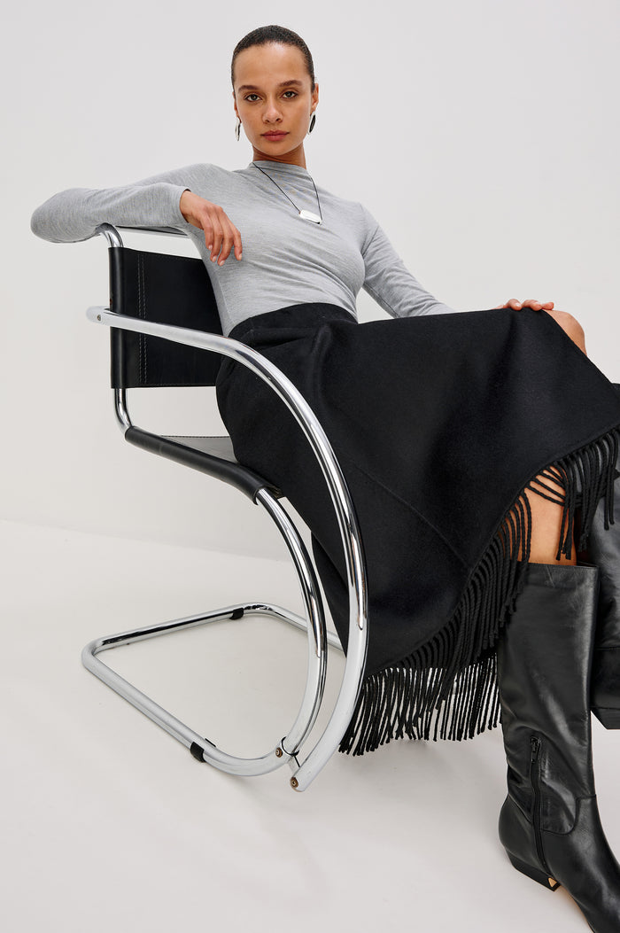 Odette Midi Skirt in Wool Blend With Wrap Design, Hidden Side Button Closure, and Fringe Hem in Black Colorway - Full Body Front View Featured on Model Sitting